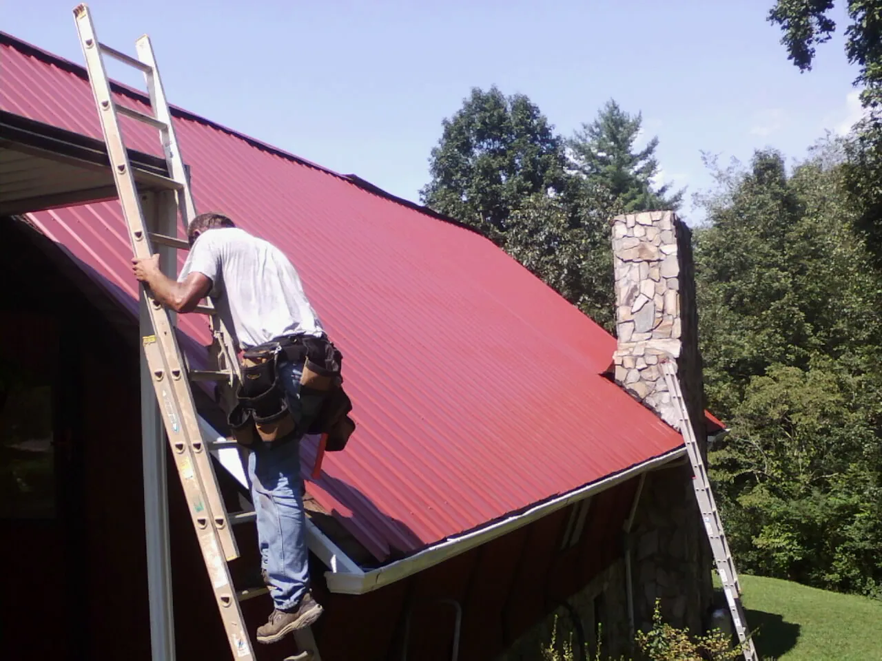 Roofer on a ladder beside a red metal roof and stone chimney — Soffit & Fascia Repair by Roofing Brothers in Assonet
